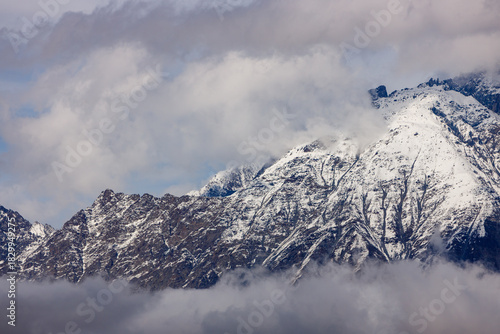 Snow-capped, jagged mountains rise above swirling clouds under a partly cloudy sky, with soft diffused lighting highlighting rocky textures and white snowfields.