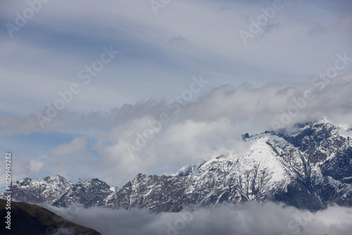 Snow-capped mountains rise above swirling clouds under a soft, overcast sky, with muted blues and grays dominating the dramatic, misty landscape.