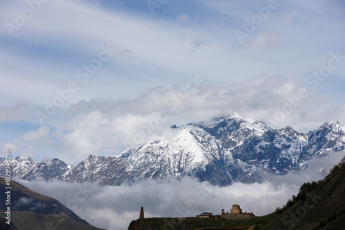 Snow-capped mountains rise above a sea of clouds, with a stone church and tower perched on a green hillside under a partly cloudy sky.