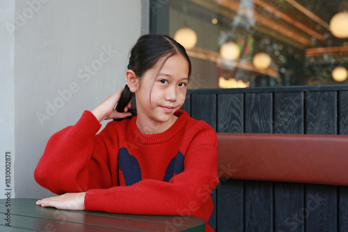 Portrait of charming Asian young girl in red sweater sitting at a table in a cafe, posing while gently touching her hair and looking directly at camera.