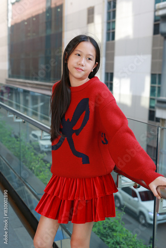 Portrait of stylish Asian young girl in red sweater and skirt, standing and leaning on a railing on an urban skywalk or balcony, looking away with a thoughtful expression.