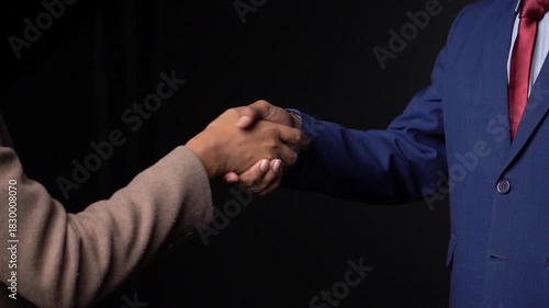 A handshake between two people one wearing a tan jacket and the other wearing a blue suit with a red tie against a dark background