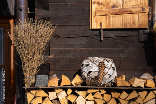 Stack of firewood logs on a shelf against a dark wooden cabin wall, featuring a decorative bundle of twigs and a woven natural sphere. Concept of cozy home, winter, and rustic lifestyle.