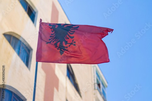 Albanian red and black national flag waving in city street, building and sky background. Symbol of patriotism, pride, tourism, and travel in Europe