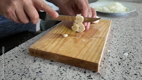 A person's hands cut slices of garlic with a knife on a wooden cutting board.