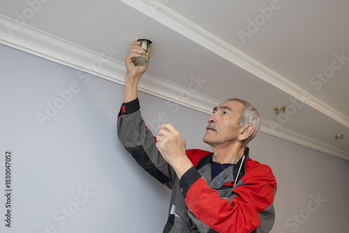 An elderly electrician changes a light bulb on the ceiling and installs a new lamp.