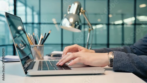 Close up of hands typing on a modern laptop keyboard in a bright office with a desk lamp and
