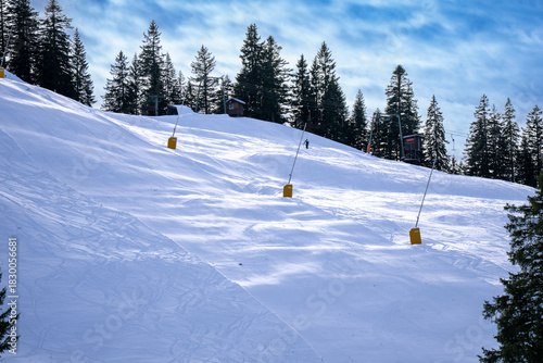 A wide, snowy ski slope on a mountain, showing clear ski tracks, forest on the ridge, and part of a ski lift. Depicts winter sports, recreation, and mountain tourism under a blue sky.
