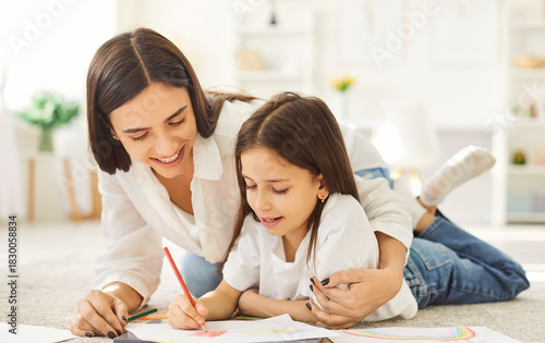 Happy mother and child drawing a picture together. Smiling little girl together with mom lying on the floor at home and drawing with colored pencils on paper. Family leisure concept