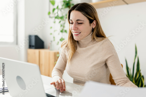 Young adult woman working on business documents at home office. Smiling professional woman multitasking with papers and laptop. Freelance productivity and digital lifestyle concept.