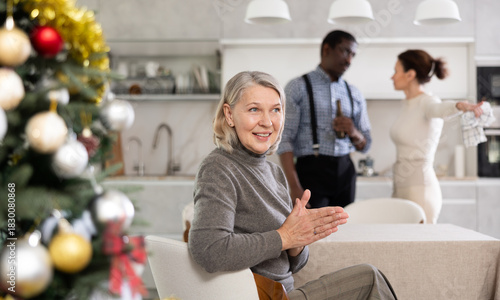 Glad old woman sitting at the kitchen table next to Christmas-tree with her back to middle-aged man and woman quarreling together
