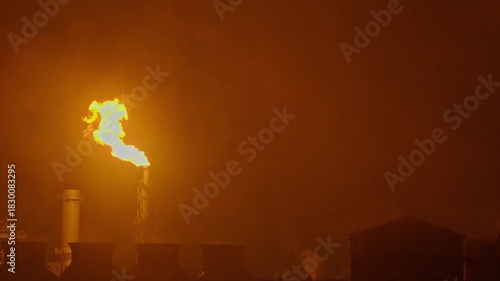 An industrial flare stack burns off excess gas with a large, bright orange flame and thick smoke against a dark night sky, highlighting energy production and potential environmental impact.