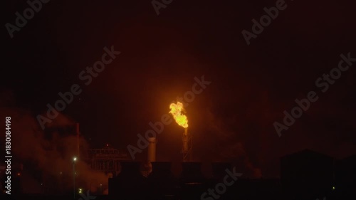 An industrial flare stack burns off excess gas with a large, bright orange flame and thick smoke against a dark night sky, highlighting energy production and potential environmental impact.