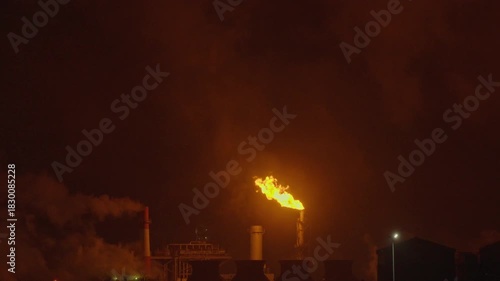 An industrial flare stack burns off excess gas with a large, bright orange flame and thick smoke against a dark night sky. Slow motion footage.