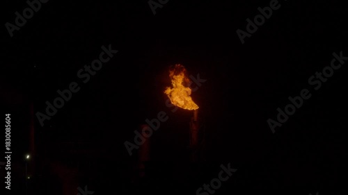 An industrial flare stack burns off excess gas with a large, bright orange flame and thick smoke against a dark night sky. Slow motion footage.