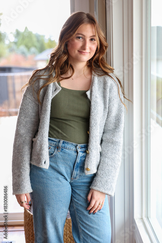 Pretty young woman wearing a soft, comfortable sweater and jeans standing near a window with a friendly smile on her face. 