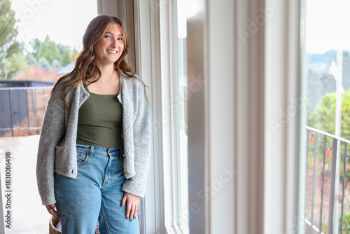 Pretty young woman wearing a soft, comfortable sweater and jeans standing near a window with a friendly smile on her face. 