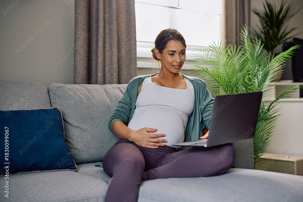 Naklejka premium Smiling pregnant woman using laptop while relaxing on sofa at home.
