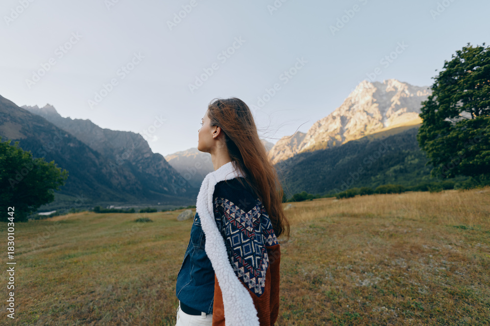 Fototapeta premium Woman in a meadow looks toward mountains in a wide landscape, wearing a patterned shawl; travel portrait of nature, solitude and serene outdoor exploration in golden light.