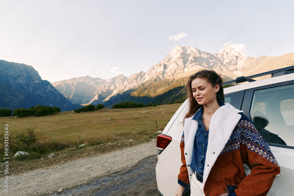 Fototapeta premium Woman by car on mountain road during outdoor travel, scenic landscape and nature view. Young female in warm jacket stands beside vehicle with peaks in background and calm expression
