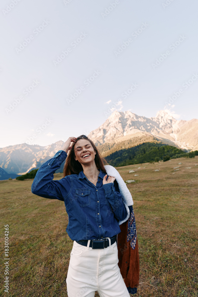Fototapeta premium Woman with smile in denim shirt and white pants standing in mountains meadow, holding a jacket over shoulder. Outdoors portrait with natural light, alpine landscape and relaxed mood.