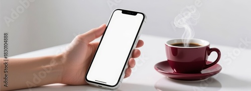 Hand holding smartphone with blank screen beside steaming coffee cup on white table