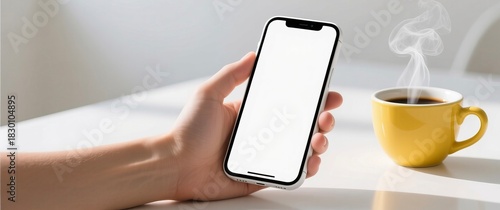 Hand holding smartphone with blank screen beside steaming yellow coffee cup on white table