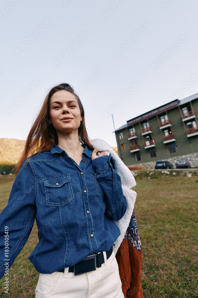 Fototapeta premium Woman smiling in denim shirt holding a jacket outdoors in a grassy field near a residential building, casual portrait with relaxed pose, natural light and confident expression.