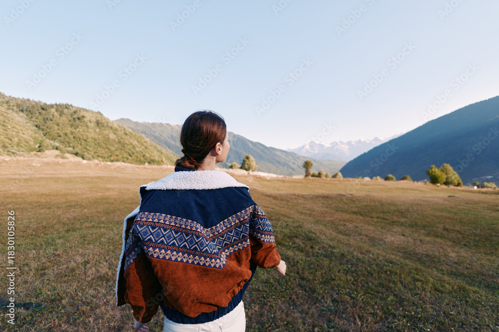 Fototapeta premium Woman in a cozy sweater walking across a mountain meadow, nature landscape with scenic outdoors scenery. Back view portrait captures calm hiking journey and tranquil horizon.