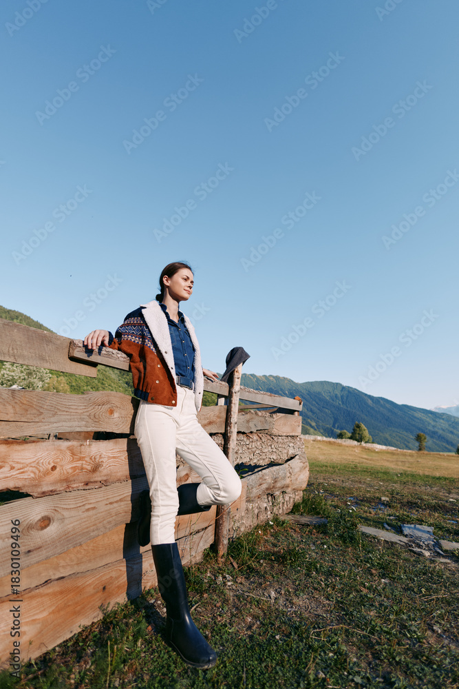 Fototapeta premium Woman leaning on wooden fence in mountains countryside meadow under clear blue sky, wearing jacket and boots, full body portrait in rural outdoors setting for fashion and travel.