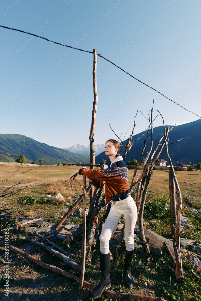 Fototapeta premium Woman countryside fence sticks mountains field: Young woman leans on rustic stick fence in open field with mountain valley backdrop, wearing sweater and boots for rural outdoor lifestyle.