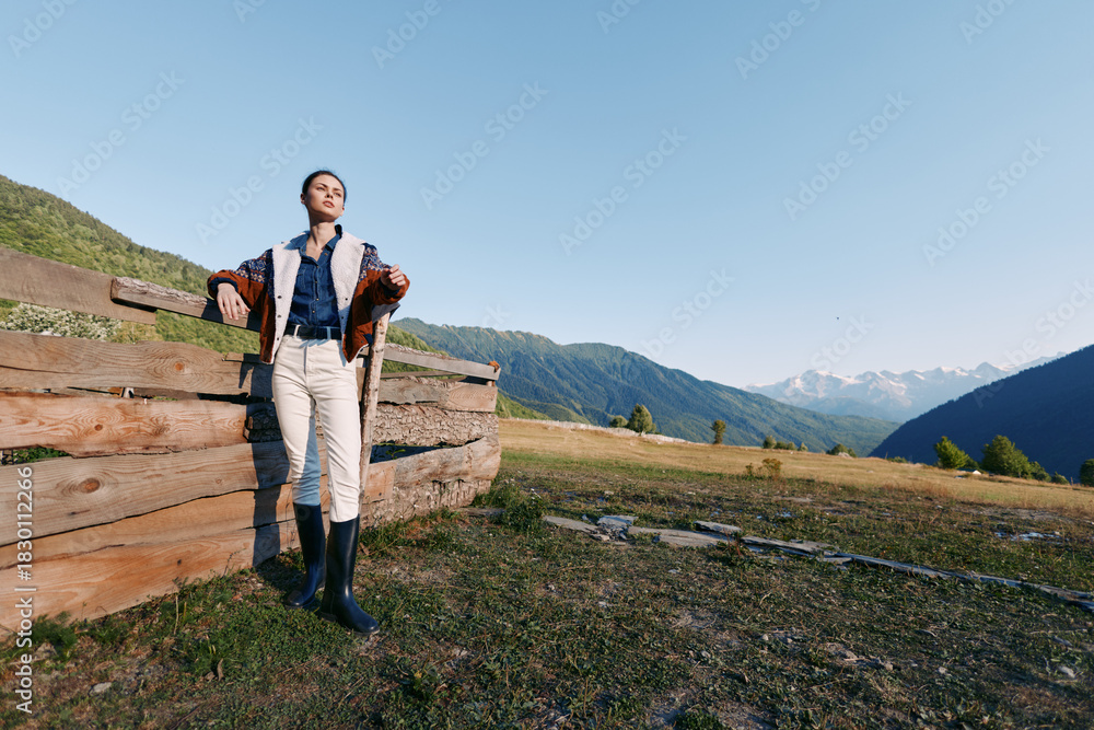 Fototapeta premium Woman leans on wooden fence in alpine meadow against distant mountains, countryside travel scene. Casual outfit with boots, relaxed portrait in wide open landscape under clear blue sky.