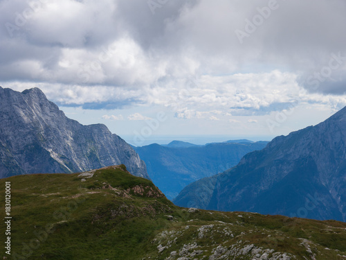 A panoramic alpine landscape of Montgart in the Alps featuring rugged rocky cliffs, soft green slopes, a winding mountain road and a dramatic cloudy sky