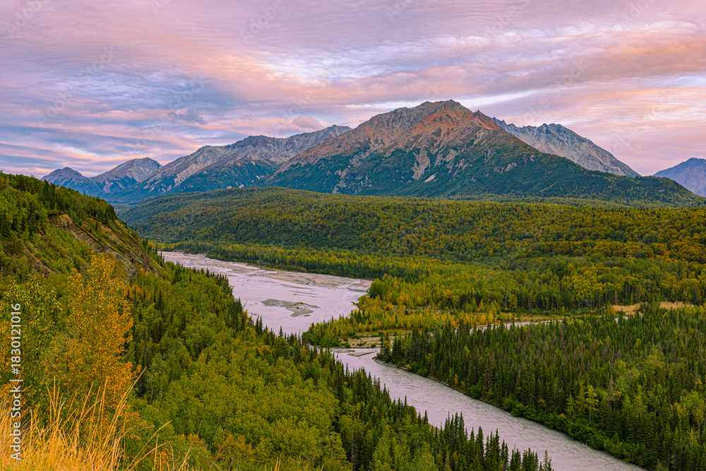 Fototapeta premium A winding glacial river cuts through dense boreal forest at golden hour along the Glenn Highway from Valdez to Anchorage, with sunset-lit Chugach Mountains glowing under pastel skies in early fall.