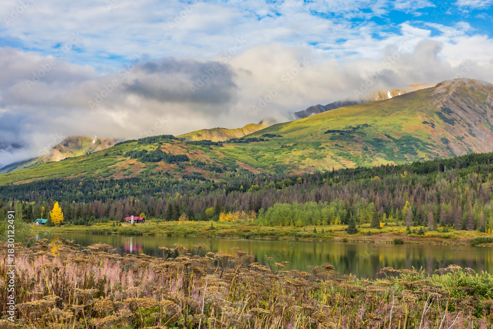Naklejka premium A peaceful autumn lake along the Seward Highway (AK-1 south to Seward), featuring a red cabin and lone golden birch reflected in calm waters, backed by mist-shrouded Chugach Mountains under dramatic c