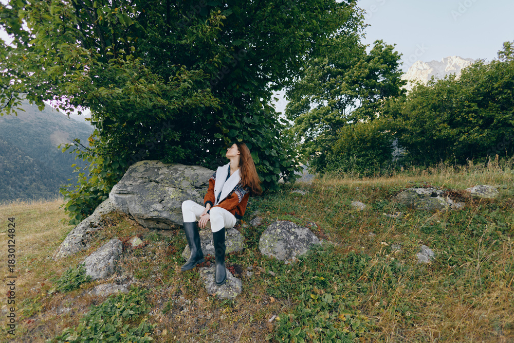 Naklejka premium Woman sitting on rock in mountain meadow under trees, looking up relaxed. Outdoor portrait with boots and coat, natural landscape, greenery and rocky foreground and sunlight.