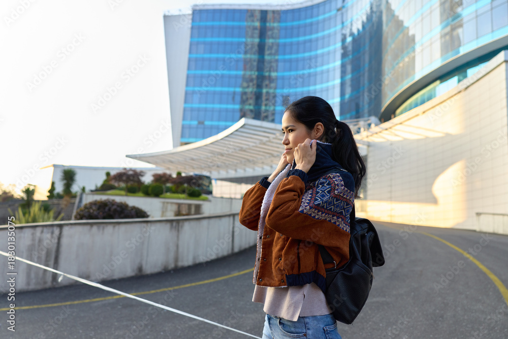 Obraz premium Woman street backpack jacket city outdoor urban phone casual architecture talking on her cellphone beside modern buildings during daylight, showcasing travel, communication and city life