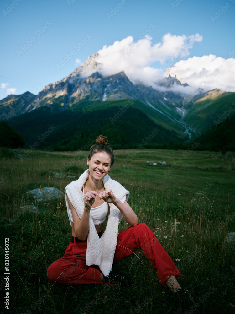 Naklejka premium Woman in red pants sits in a meadow with mountains behind, smiling and relaxed, enjoying outdoor nature, casual pose, scenic landscape for travel, wellness and hiking