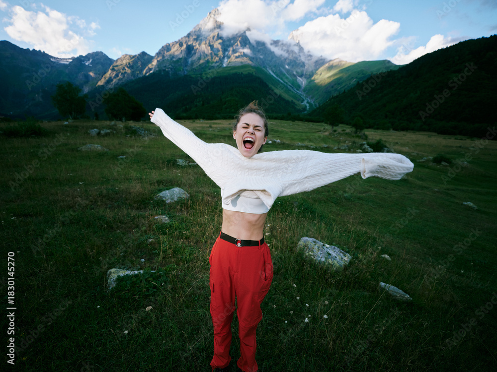 Obraz premium Outdoor celebration in alpine landscape, a joyful figure spans arms wide, wearing a white sweater and red pants, with mountains and green valley in the background