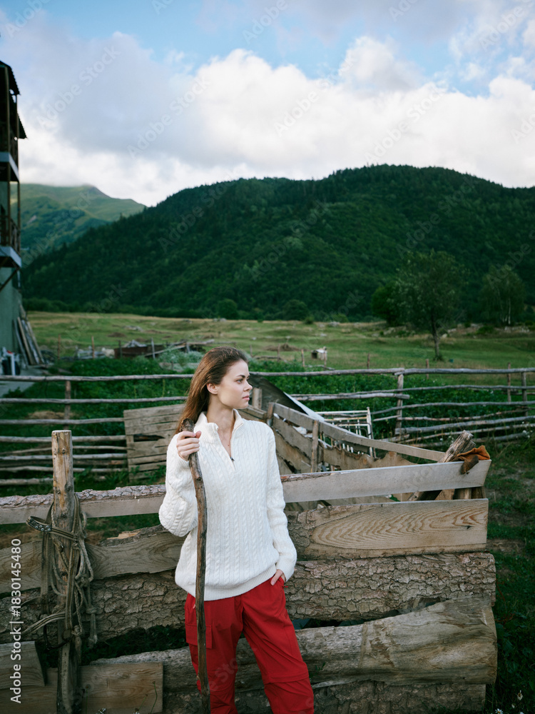 Naklejka premium Woman, countryside, outdoor, rural, standing beside a weathered fence with a staff, white sweater and red pants, expansive meadow and distant mountains under a bright sky