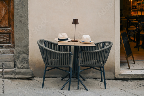 Fototapeta Naklejka Na Ścianę i Meble -  An Italian street café features a small wooden table with a lamp and two decorative hats, creating a warm and inviting outdoor scene.