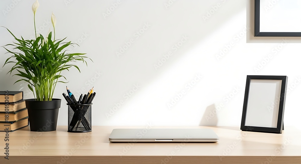 Fototapeta premium Minimalist workspace featuring a closed laptop, plant, books, picture frame, and pen holder on a wooden desk against a white wall with sunlight.