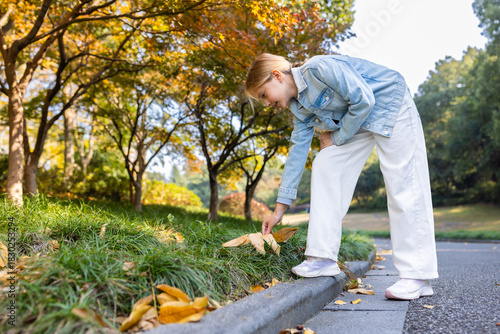 Young girl picking up fallen leaves on a park path during autumn