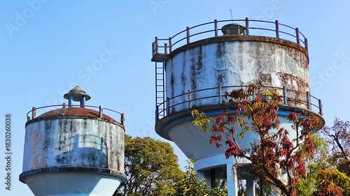 Two old water tanks rise above leafy trees under a clear sky, showing aging infrastructure and the industrial character of a small town environment.