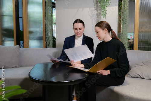 Two young professional business woman, accountant review budget or balance sheet, two busy female employees working together. Holding Document file and discussing data in paper work planning project.