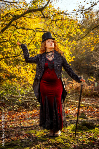 A beautiful woman with long red hair, dressed and posing in a Black top hat and tails, posing with a black cane, set against  autumn leaves and a woodland background