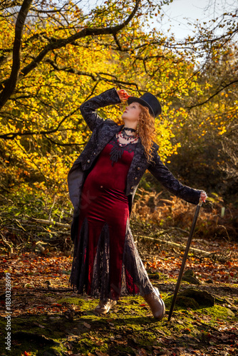 A beautiful woman with long red hair, dressed and posing in a Black top hat and tails, posing with a black cane, set against  autumn leaves and a woodland background
