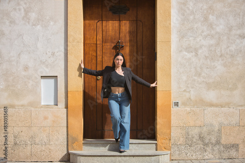 Young woman, pretty, brunette, blue eyes, Hispanic, wearing a black jacket and top, jeans, posing with her hands resting on the frame of a beautiful door. Concept: model, beauty, fashion, trendy.