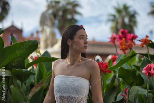 Young woman, beautiful, brunette, blue eyes, Hispanic, wearing a white top, posing while gazing into the distance surrounded by plants with pretty colorful flowers. Concept: model, beauty, fashion.