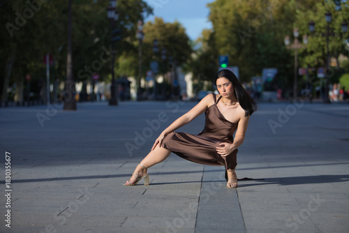 Young woman, beautiful, brunette, blue eyes, Hispanic, wearing an elegant brown chiffon dress and gold heels, posing crouched on a city sidewalk while the wind blows her hair. Concept: model, beauty.